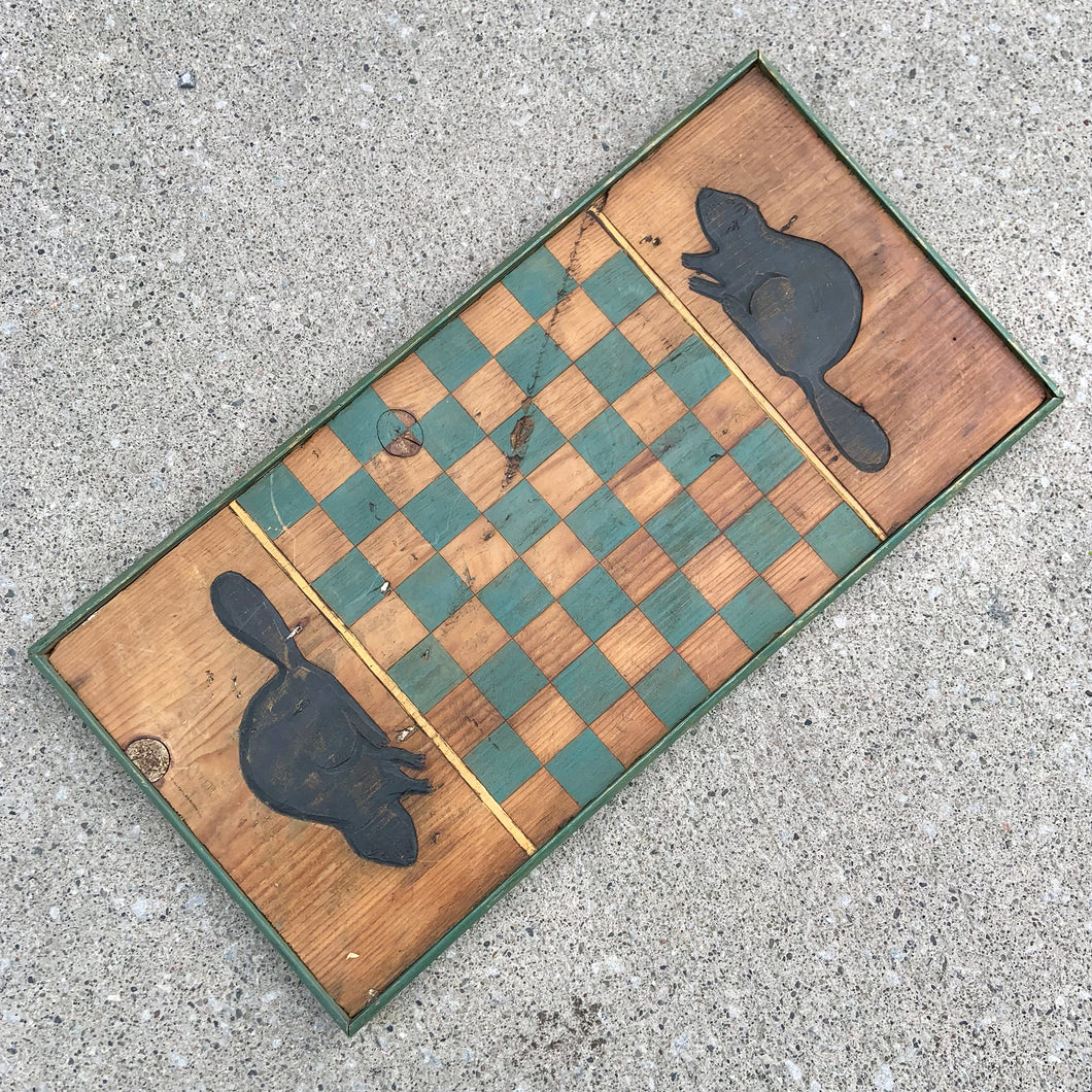 This vintage handmade primitive pine wood checker chess board features a natural, green and black stain, adorned with hand carved beavers at either end. It's a great addition to family game nights at the cottage, or as a rustic wall hanging to add a touch of nature-inspired charm to your decor.

In good vintage condition. Wired on the back for wall hanging.

Measures 12 1/2 x 24 3/4 x 1 1/4 inches