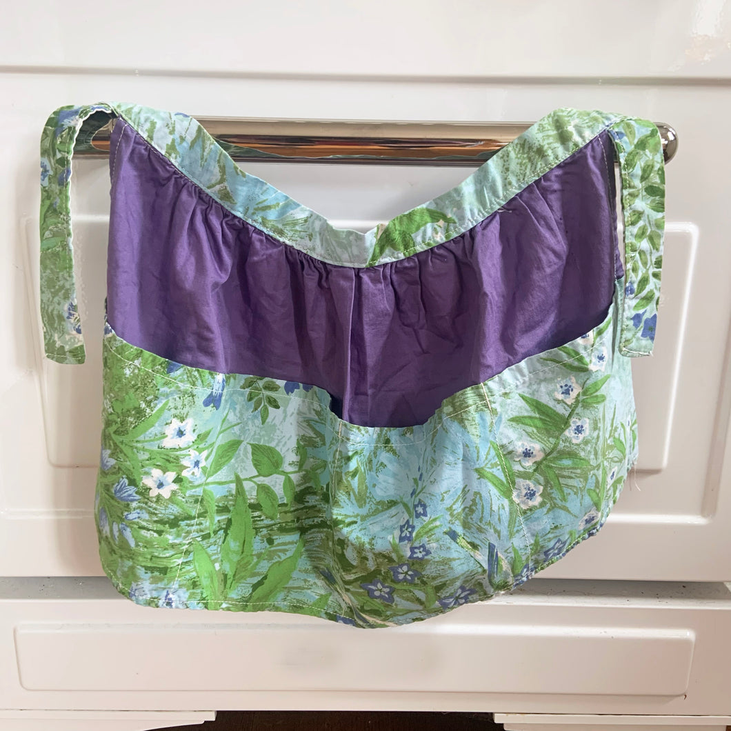 Beautifully hand made vintage fabric apron with pockets. The gathered body of the apron is made in purple fabric with the band and pockets in a pretty robin's egg blue with white and green flowers.  In excellent condition, free from rips/stains.  The band measures 42 inches, body measures 25 1/2 x 13 inches