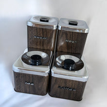Load image into Gallery viewer, Vintage &quot;The Queen Line&quot; four piece metal kitchen canister set featuring faux brown wood grain, chrome lids with black plastic handles, in two sizes. Each has period style typography in black plastic letters overpainted in silver for flour, sugar, coffee and tea. Stamped on the bottom with, The Queen Line, By A.R Lite, Made in/Fabrique au Canada.
