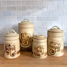 Load image into Gallery viewer, Add a touch of retro charm to your kitchen with this Vintage Retro Kitchen Stoneware Canister Set by Pottery Craft, USA. The set includes canisters for flour, sugar, coffee and tea, each featuring a unique speckled beige and brown design with the name in relief, glazed in a contrasting rusty brown. The funky stylized font and handy handles on the lids add to its appeal. Crafted in the 1970s, this set is a must-have for any retro kitchen lover!

The flour, sugar tea in excellent condition, coffee tiny chip
