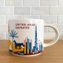 Load image into Gallery viewer, Perfect for your morning cuppa joe to make you feel like a world traveller! This fabulous collectible UNITED ARAB EMIRATES ceramic mug was produced by Starbucks as part of their &quot;YOU ARE HERE&quot; collection, 2018. A white ceramic mug printed design in shades of pink, blue, red and yellow with blue interior. Celebrate the Make it Happen country!

In excellent condition, free from chips/cracks/wear.

Measures 3 5/8 x 3 3/8 inches

Capacity 14 ounces
