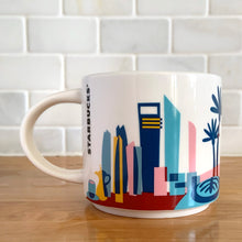 Load image into Gallery viewer, Perfect for your morning cuppa joe to make you feel like a world traveller! This fabulous collectible UNITED ARAB EMIRATES ceramic mug was produced by Starbucks as part of their &quot;YOU ARE HERE&quot; collection, 2018. A white ceramic mug printed design in shades of pink, blue, red and yellow with blue interior. Celebrate the Make it Happen country!

In excellent condition, free from chips/cracks/wear.

Measures 3 5/8 x 3 3/8 inches

Capacity 14 ounces
