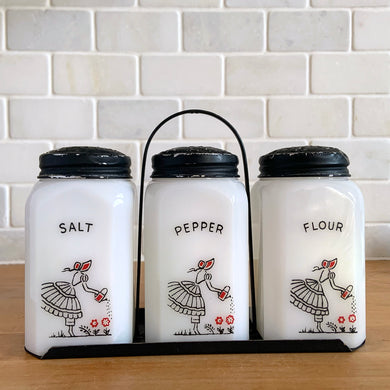 Vintage Tipp City trio of milk glass shakers for salt, pepper and flour in metal stand featuring a printed design of a girl watering flowers. The glass has a slightest glow under black light, likely due to manganese in the glass. Crafted by McKee Glass, 1940s. Beautiful art deco tableware to add to your shaker collection.

In vintage condition, no chips, print is intact and paint on metal lids has age-related wear.

Each shaker measures 2 3/8 x 2 3/8 x 4 1/2 inches