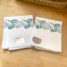 Load image into Gallery viewer, A lovely pair of vintage white 100% percale cotton pillowcases, hand embroidered with a blue daisy flowers, graphic blue leaves. These new old stock linens will be a gorgeous addition to your bedding!

In excellent condition, free from tears. Age-related yellowing at the bands. Original stickers, made in Japan.

Measures 30 3/4 x 18 inches
