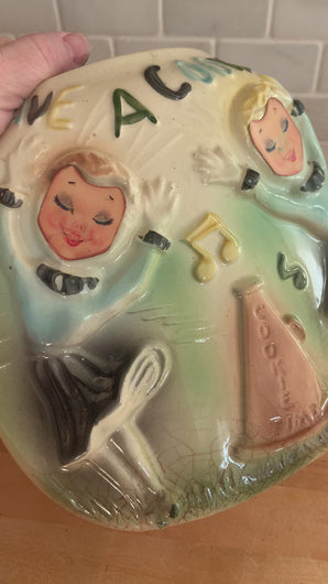 Movie of rare vintage ceramic corner cookie jar features brunette and blonde cheerleaders in old-fashioned outfits, cheering underneath the words Have A Cookie in between is a megaphone on the grass that reads cookie time all glazed in shades of green, blue, yellow brown and black on a cream ground, topped with a rust-brown megaphone on the lid. The piece-de-resistance are the cheerleaders holographic facial expressions that change from eyes open with a wide smile to eyes shut with closed smiles. 
