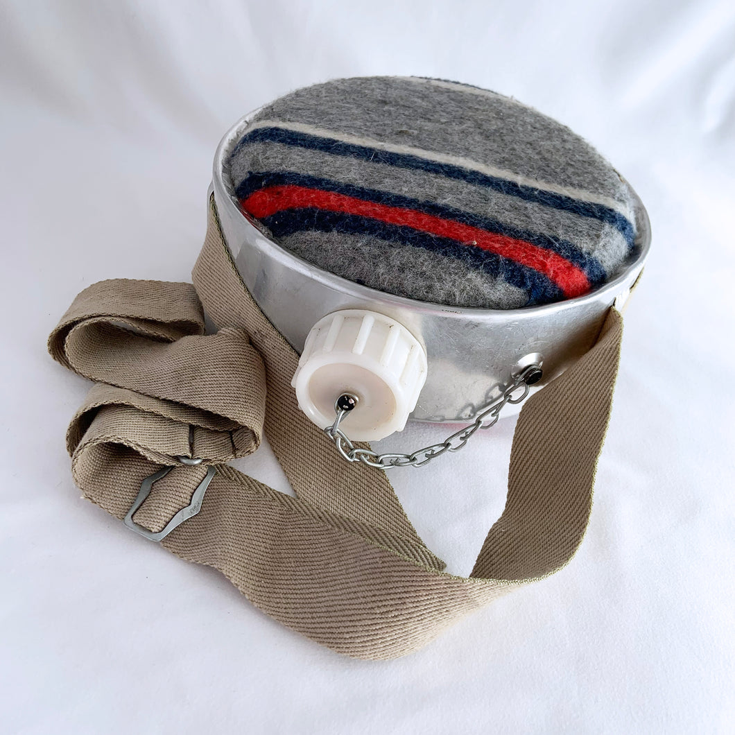 Bring back memories of childhood camping or hiking trips with this vintage canteen! It has a round aluminum body with a wool blanket wrap to keep liquids cool and is easily carried by the canvas strap. The twist plastic cap keeps everything contained and it's chained to the canteen so there's no chance of it being misplaced. Great for regular use or for prop styling. Made in Korea, circa 1960s.   In excellent used vintage condition.    Measures 7-1/4