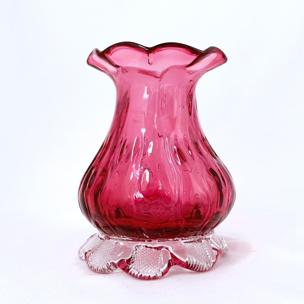Beautiful, heavy-weighted cranberry glass vase with clear scalloped foot. The vase has an urn-shape with a ribbed detail and ruffled edge. A very nice piece of glass to use for a floral bouquet or repurpose to fold make-up brushes or q-tips. We see a lot of these pieces attributed to Pilgrim Glass, but without a maker's mark we can't be certain.  In excellent condition, free from chips/cracks/repairs.  Measures 2 7/8 x 4 inches