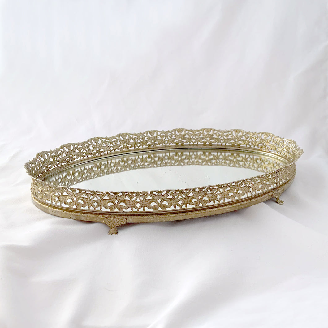 This elegant vintage Hollywood Regency style oval-shaped mirrored dresser vanity tray, featuring brass ormolu details, is perfect for displaying perfume bottles, trinket dishes, and makeup. Alternately, hang it on the wall to add glitz and glamour to your décor.  In excellent condition.  Measures 12 x 9 x 1 1/4 inches