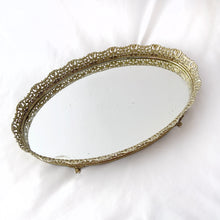 Load image into Gallery viewer, This elegant vintage Hollywood Regency style oval-shaped mirrored dresser vanity tray, featuring brass ormolu details, is perfect for displaying perfume bottles, trinket dishes, and makeup. Alternately, hang it on the wall to add glitz and glamour to your décor.  In excellent condition.  Measures 12 x 9 x 1 1/4 inches
