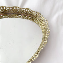 Load image into Gallery viewer, This elegant vintage Hollywood Regency style oval-shaped mirrored dresser vanity tray, featuring brass ormolu details, is perfect for displaying perfume bottles, trinket dishes, and makeup. Alternately, hang it on the wall to add glitz and glamour to your décor.  In excellent condition.  Measures 12 x 9 x 1 1/4 inches
