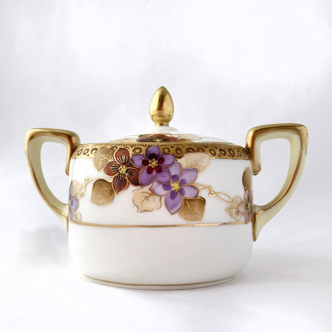 Elegant and beautifully hand painted antique porcelain oval-shaped creamer and covered sugar bowl with clematis flowers in shades of purple and gold vines with gold moriage details. The base of each piece show the Noritake stamp called the Maruki mark which was produced between 1906 to 1925. The sugar bowl is in excellent condition free from chips, cracks or repairs and has minor wear to the gold band on the lid, see photos. The creamer has a vertical crack on the body near the handle.