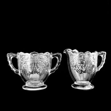 Vintage clear pressed glass creamer and sugar with silver overlay in a design of vase with florals.  In good vintage condition, free from chips/cracks. Silver is worn in a couple of places.  Creamer measures 2 7/8 x 3 3/4 inches  Sugar measures 3 3/8 x 3 3/8 inches