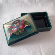 Load image into Gallery viewer, A lovely vintage &quot;Columbine&quot; dresser box with a jewel-toned flower against a green wash ground. Design by Walter Moorcroft and produced by Moorcroft Pottery, England, circa 1950.  Stamped with the WM Moorcroft signature and &quot;Made in England&quot; along with paper label having the Royal Warrant and &quot;By Appointment, Potter to the late Queen Mary&quot;.   In excellent condition, free from chips, cracks, repairs; crazing present.  Measures 4.75&quot; x 3.5&quot; x 2&quot;
